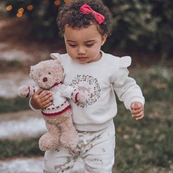 A toddler wearing a matching white long-sleeve top and pants set with a holiday wreath and deer print, holding a plush teddy bear. The child has curly hair with a pink bow and is photographed outdoors in a soft-focus natural setting.