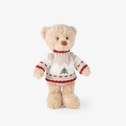 A plush teddy bear wearing a cream knit sweater with red trim and a green Christmas tree motif, photographed against a clean white background.