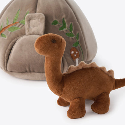 A brown plush dinosaur toy in the foreground with a beige dome-shaped soft play house featuring embroidered green leaves and a small mushroom in the background, set against a clean white studio backdrop.