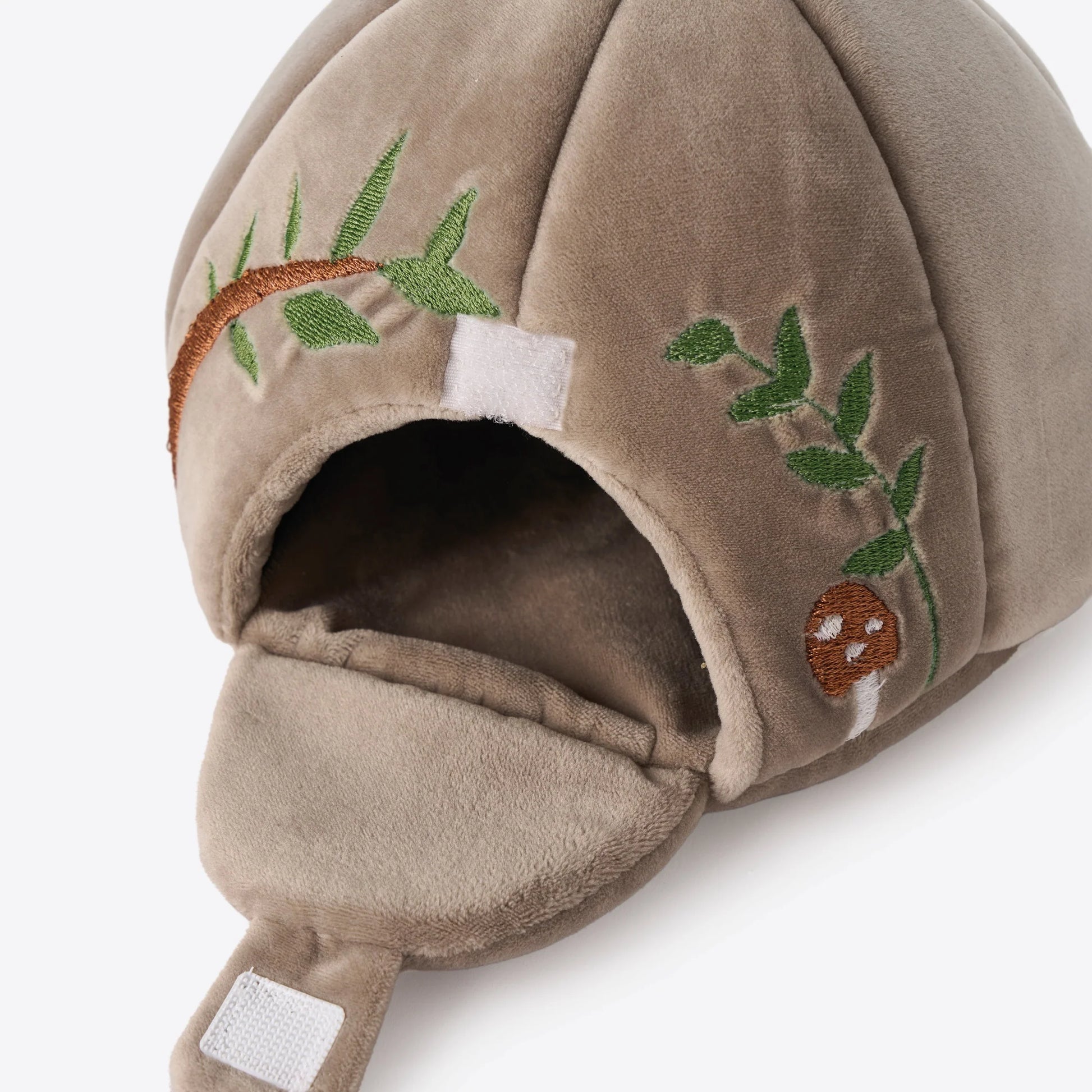 A close-up of a plush brown cave with embroidered green leaves, a brown mushroom, and a white hook-and-loop fastener on a light gray background.