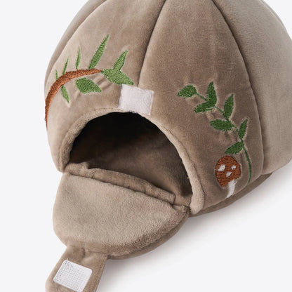 A close-up of a plush brown cave with embroidered green leaves, a brown mushroom, and a white hook-and-loop fastener on a light gray background.