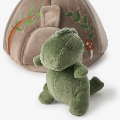 A small plush dinosaur toy in olive green sitting in front of a beige fabric play tent with embroidered green leaves and a brown mushroom.
