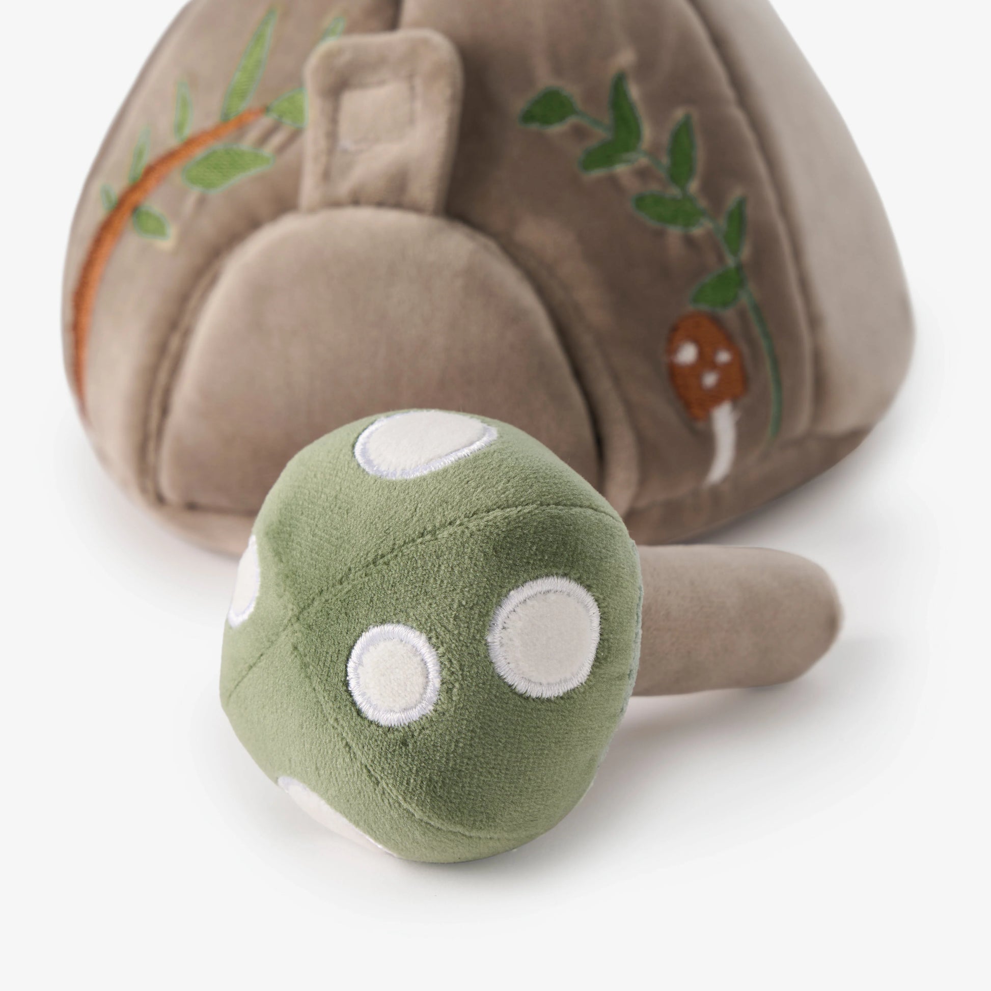 A plush mushroom toy with a green cap featuring white embroidered spots and a tan stem, shown in front of a larger brown mushroom-shaped plush with embroidered green leaves and a small brown accent on a white background.