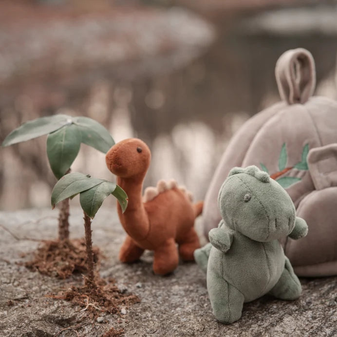 A cozy arrangement of plush toys on a stone surface: a small green dinosaur in the foreground, a smaller orange-brown dinosaur behind it, and a large taupe house with a bunny ears detail and leaf embroidery in the background. Two small potted plants sit beside them against a softly blurred outdoor-style backdrop.