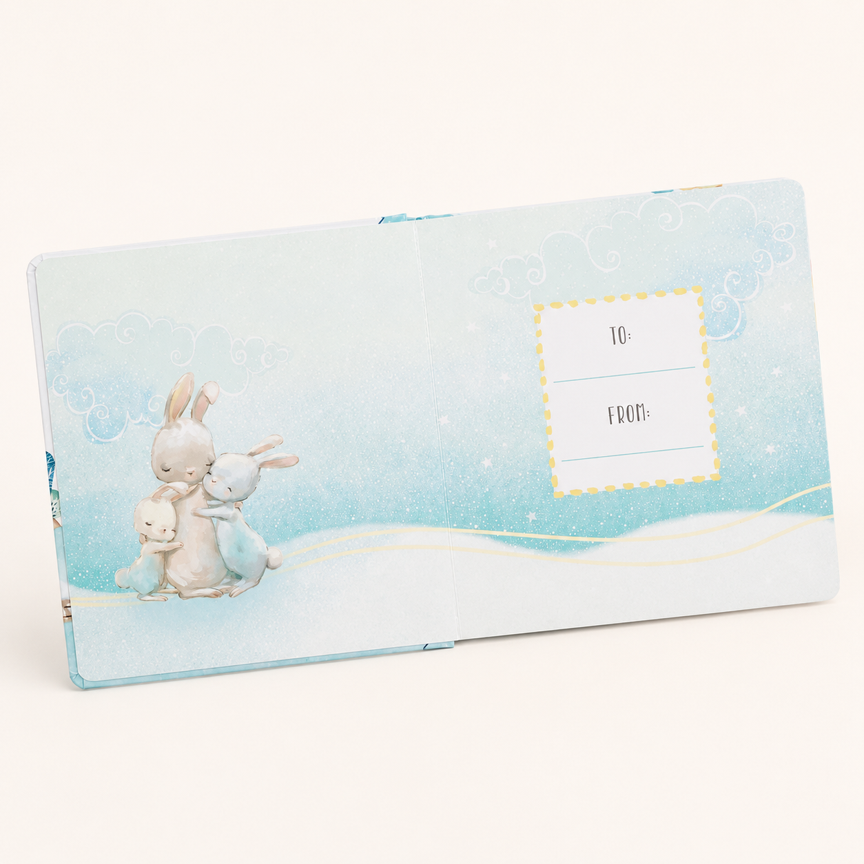 A pastel blue baby keepsake album shown open on a warm off-white background. The cover features an illustration of three rabbits in soft blue and cream tones, with a dotted yellow 'TO:' and 'FROM:' label on the right page.