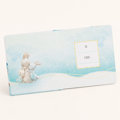 A pastel blue baby keepsake album shown open on a warm off-white background. The cover features an illustration of three rabbits in soft blue and cream tones, with a dotted yellow 'TO:' and 'FROM:' label on the right page.