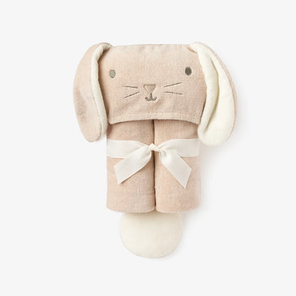 A plush bunny-shaped baby hooded bath towel in a soft blush beige color, featuring long floppy ears, embroidered facial details, and a cream-colored ribbon tied around the middle, set against a plain light gray background.