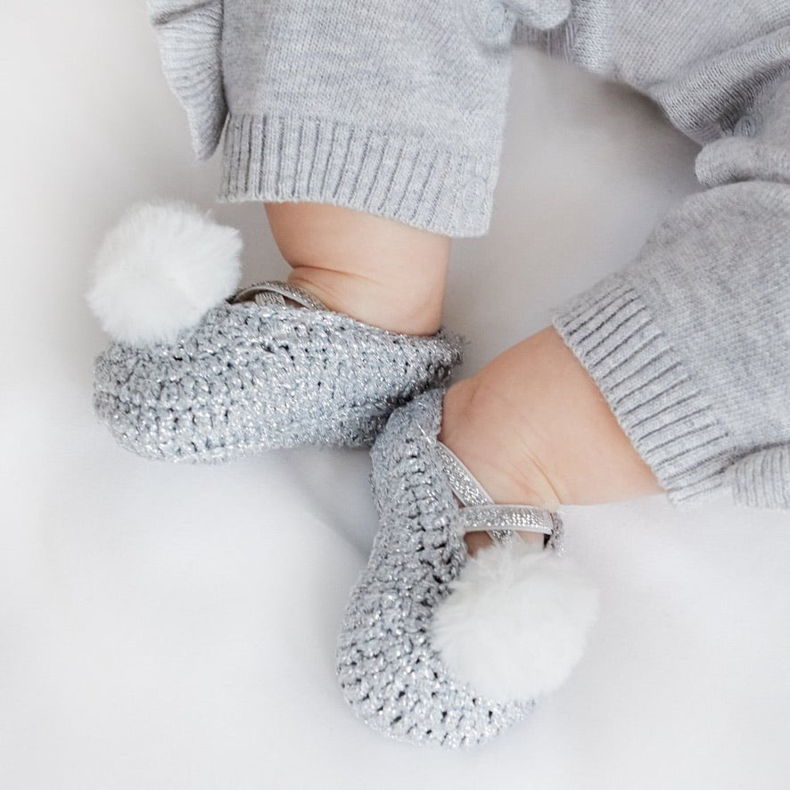 A pair of metallic gray baby booties with white pom poms, worn on a baby's feet.
