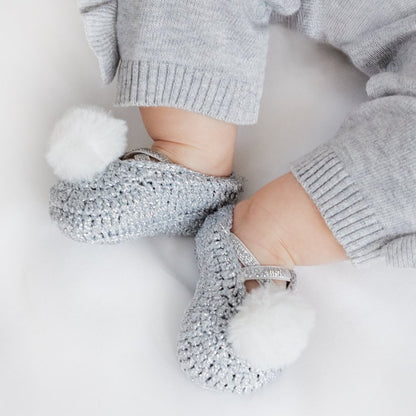 A pair of metallic gray baby booties with white pom poms, worn on a baby's feet.