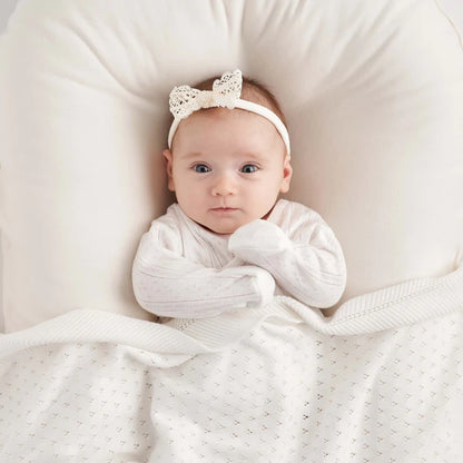 A newborn baby wearing a white four-piece matching set including a kimono wrap top, footed pants, knotted beanie, and re-usable drawstring bag.